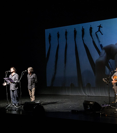 Actuación de "Su Mal Espanta" de la Cía. de Poesía La Palabra Itinerante en un escenario con músicos y proyecciones.